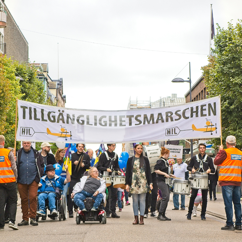Bilden visar banderollen med text Tillgänglighetsmarschen och HIL logotyp samt personer som marscherar med rullstol och utan