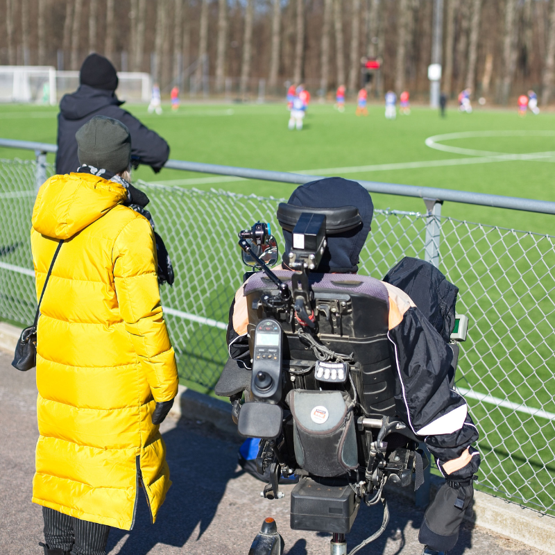 Bilden visar man i sin rullstol tittande på fotbollsmatch med sin personliga assisten i gul rock